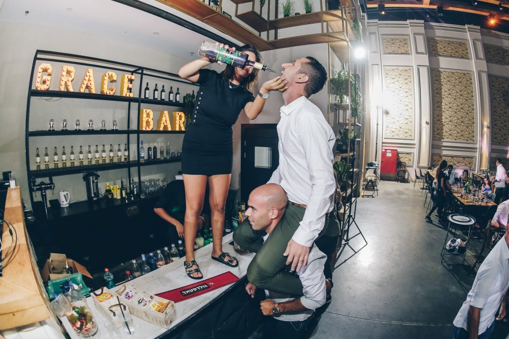 the groom drinking alcohol at a bar as he sits on his friend's shouldr while a bartender pours alcohol in his mouth at an israeli party 